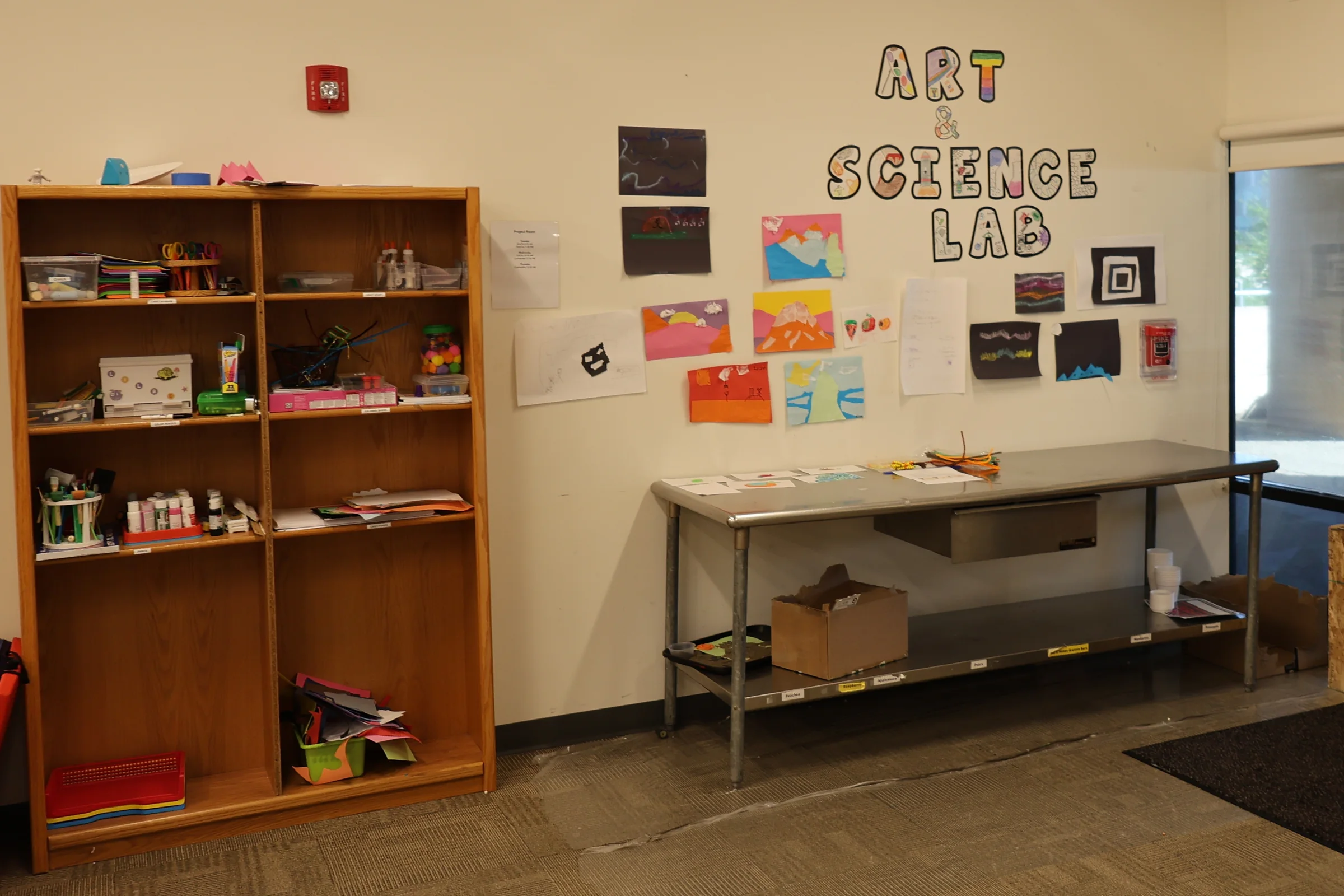 The art and science lab: a work table, supply shelving, and a wall of student artwork beneath a hand-made sign.