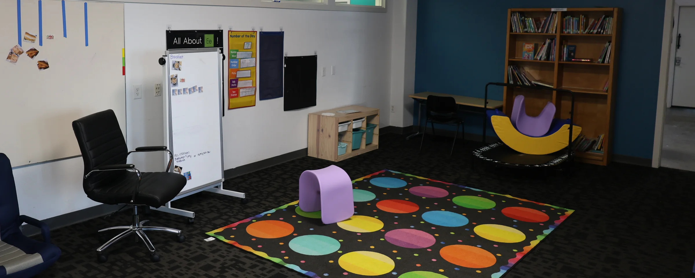 An empty Lilac Learning Center classroom: a patterned rug, a mobile whiteboard, a library shelf, a mini trampoline and a rocker.