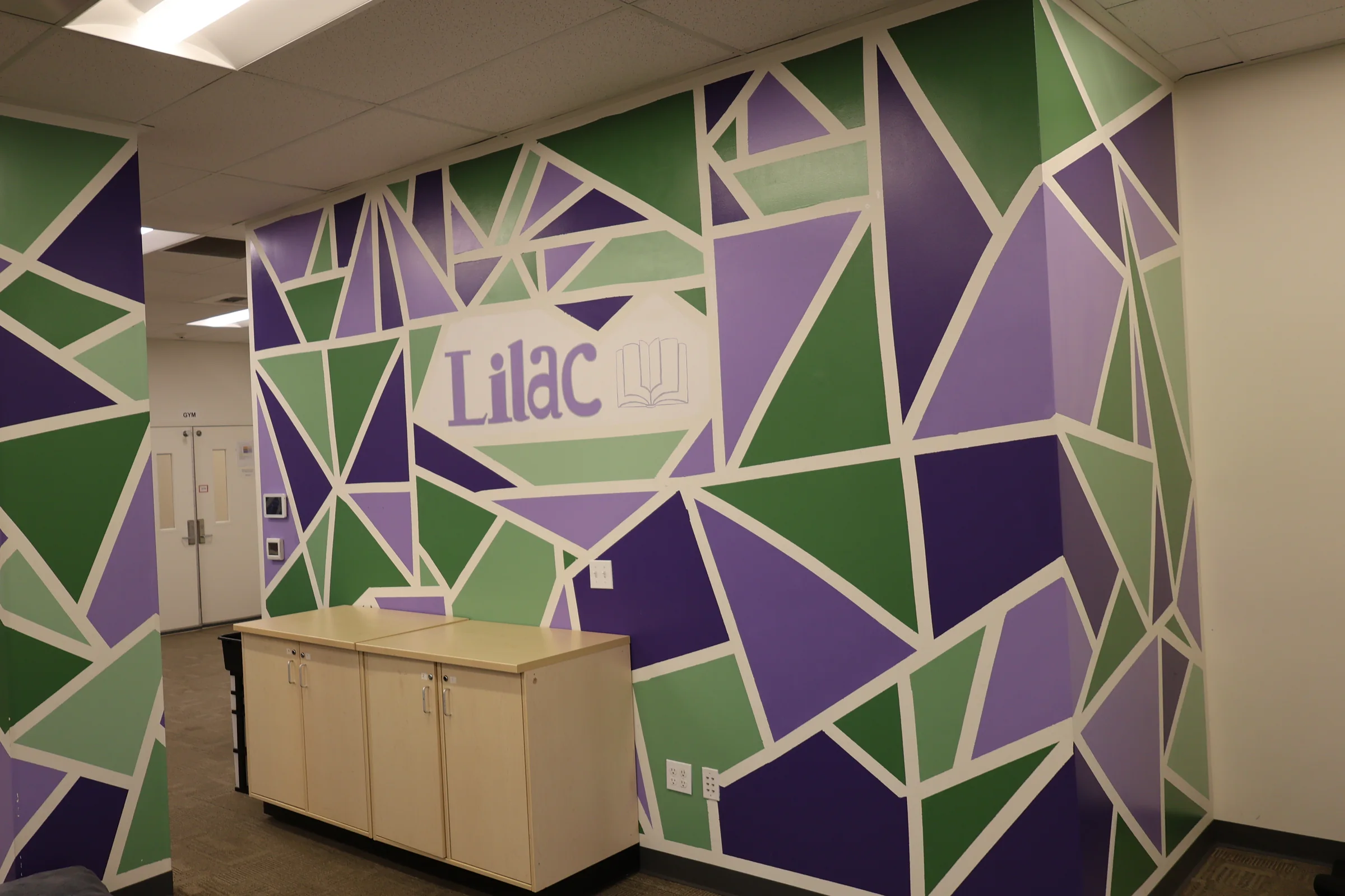 A wall painted with an angular mosaic of purple and green shapes, the Lilac Learning Center wordmark set into the middle of it.